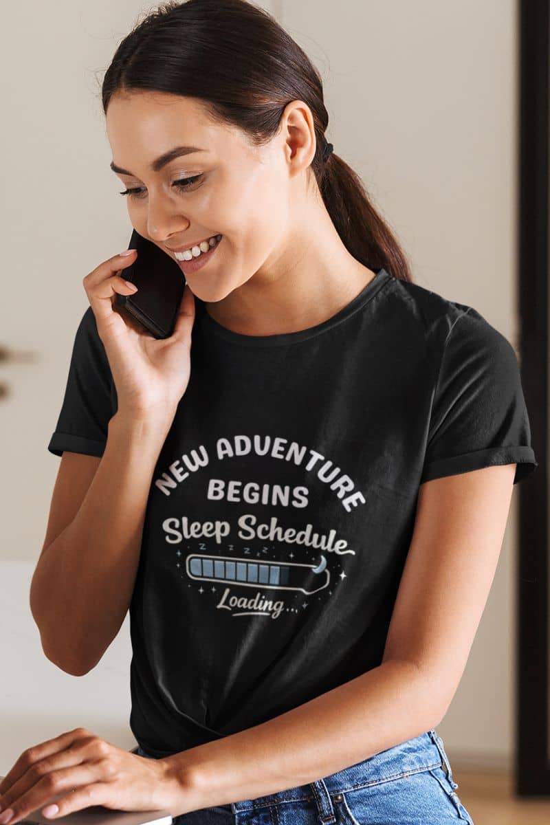 tired dad tee with a woman wearing a black t-shirt with text 'New Adventure Begins, Sleep Schedule Loading…', smiling and talking on a phone.