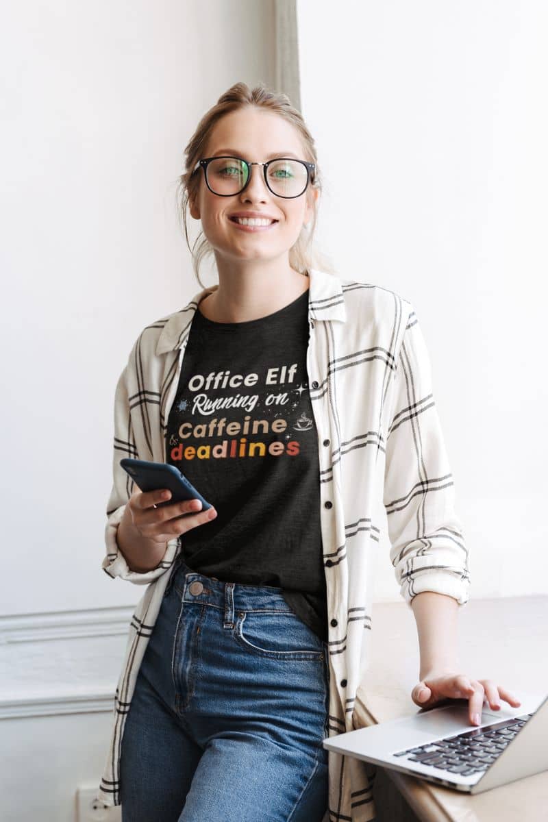 tired employee shirt with a woman wearing a black t-shirt with text 'Office Elf, Running on Caffeine & Deadlines', holding a phone and using a laptop in an office setting.