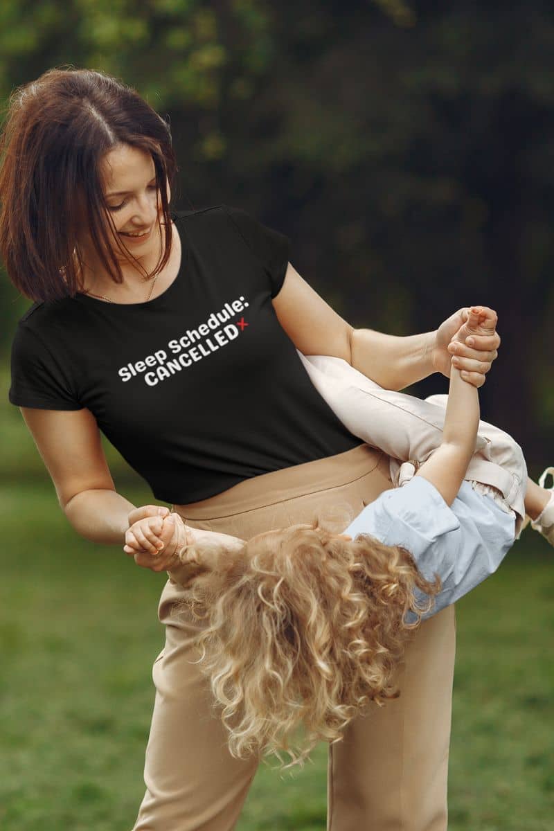 tired everyday shirt with a woman holding a child in a park wearing a black t-shirt with text 'Sleep Schedule: Cancelled'.