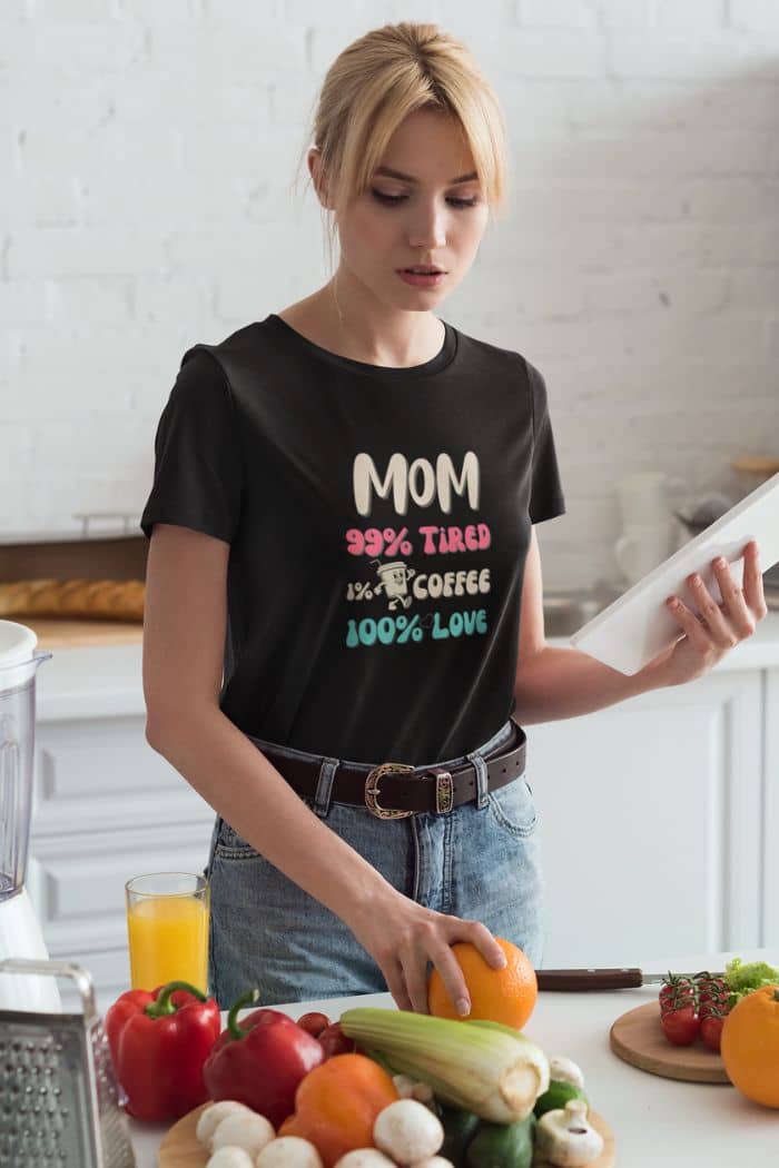 tired mom shirt with a woman in a kitchen wearing a black t-shirt with text 'Mom 99% Tired, 1% Coffee, 100% Love', surrounded by fruits and vegetables.