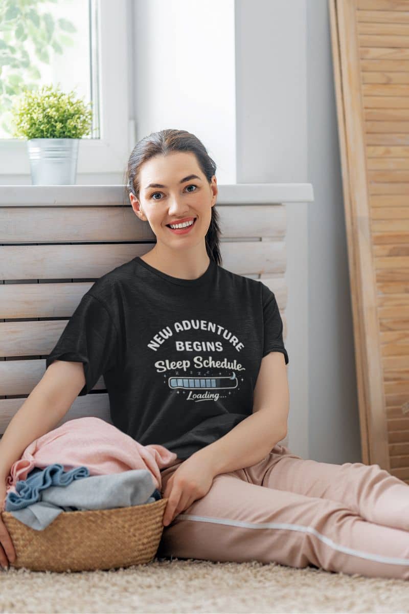 tired mom t-shirt with a woman sitting on the floor with a basket of clothes, wearing a black t-shirt with text 'New Adventure Begins, Sleep Schedule Loading…'.
