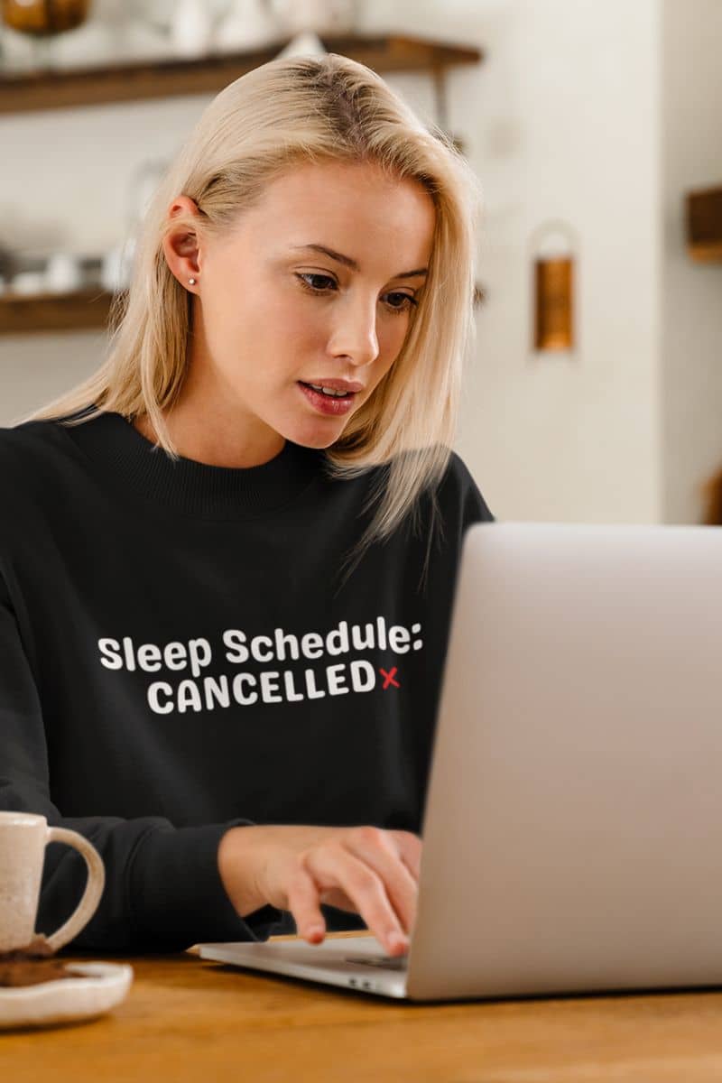 tired office sweatshirt with a person wearing a black sweatshirt with 'Sleep Schedule: CANCELLED' text, using a laptop.