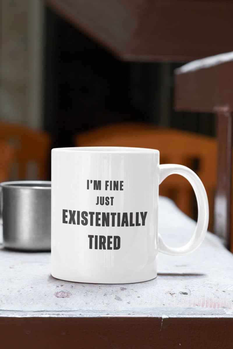 tired office worker mug with a white mug with text 'I'm fine just existentially tired' on a table.