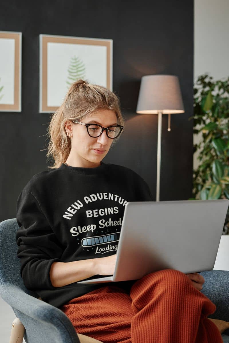 tired sweatshirt with a person wearing a black sweatshirt with text 'New Adventure Begins, Sleep Schedule Loading…', sitting on a couch using a laptop with a dark wall and lamp in the background