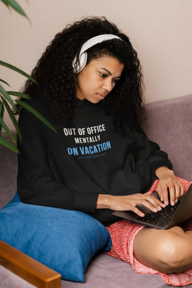 travel lover hoodie with a woman wearing a black sweatshirt with text 'Out of Office: Mentally on Vacation', using a laptop on a couch.