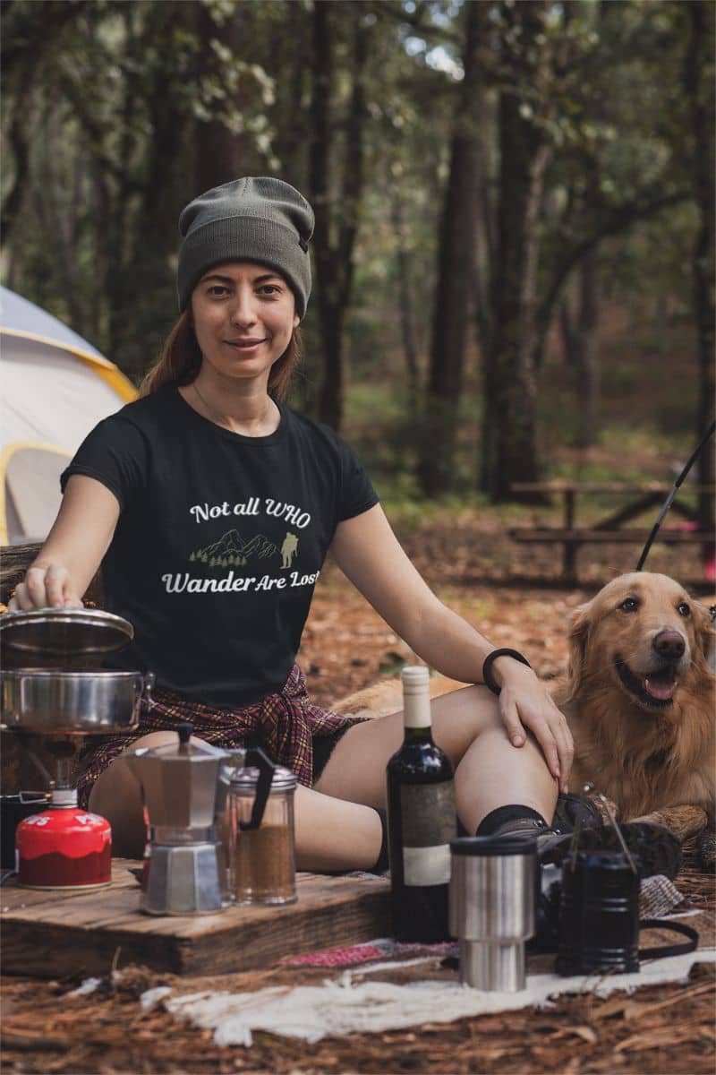travel quote shirt with a woman wearing a black t-shirt featuring the text 'Not all who wander are lost', sitting outdoors with a dog, surrounded by camping gear and drinks