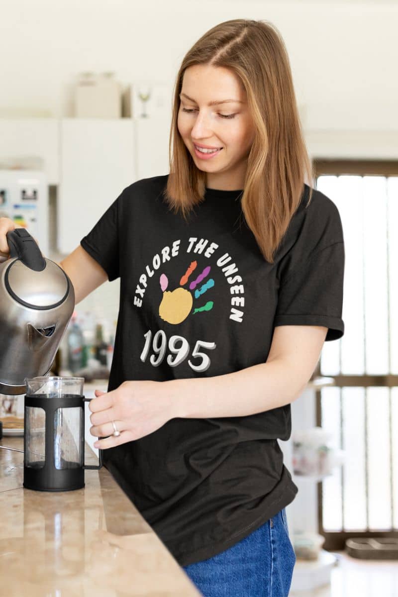 travel quote t-shirt with a woman in a kitchen wearing a black t-shirt with 'Explore the Unseen 1995' design.