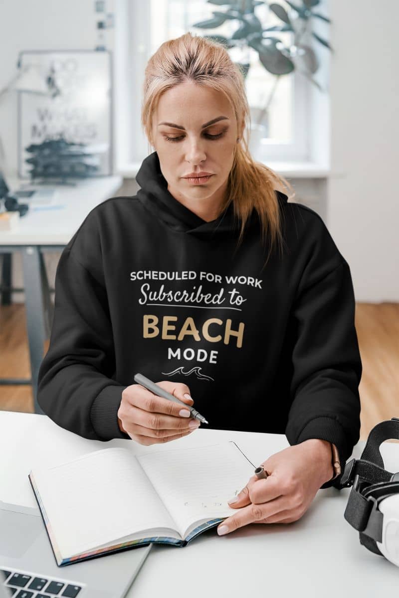travel sweatshirt with a person wearing a black hoodie with text 'Scheduled for Work. Subscribed to Beach', sitting at a desk with a notebook and pen.
