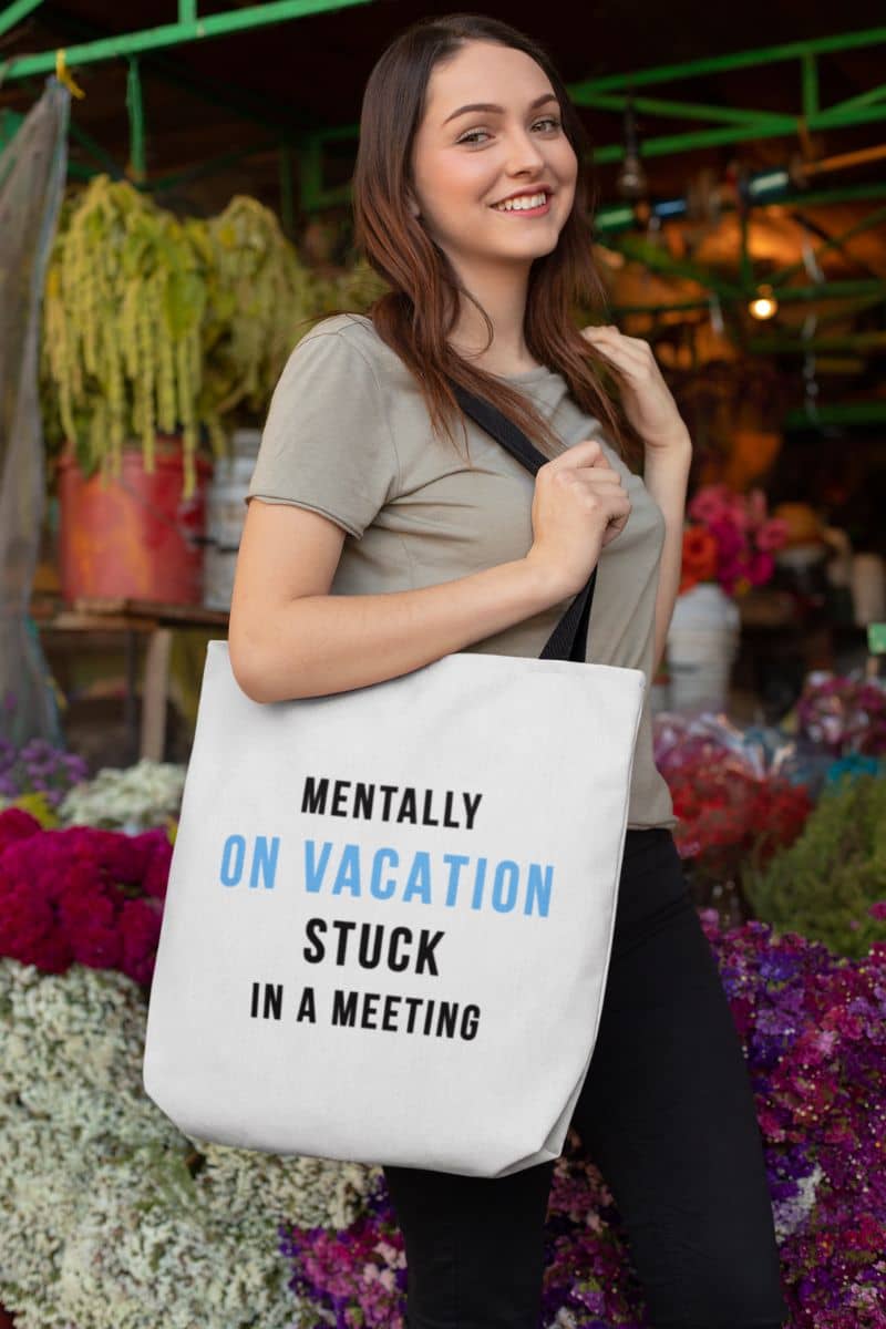 travel tote bag with a woman holding a tote bag featuring the text 'Mentally on Vacation. Stuck in A Meeting' at an outdoor market.