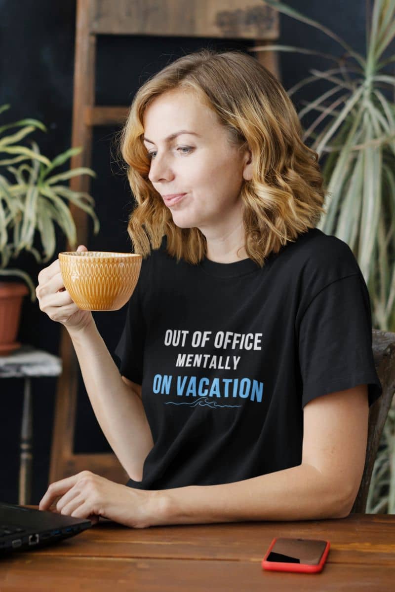 vacation lover shirt with a woman holding a mug with text 'Out of Office: Mentally on Vacation' on her shirt in a casual setting