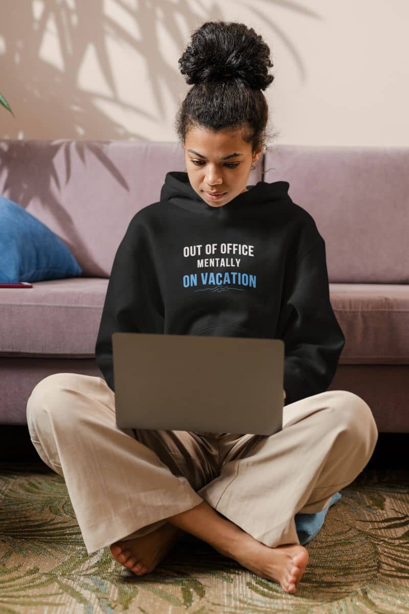 vacation sweatshirt with a person wearing a hoodie with text 'Out of Office: Mentally on Vacation' sitting on the floor using a laptop.