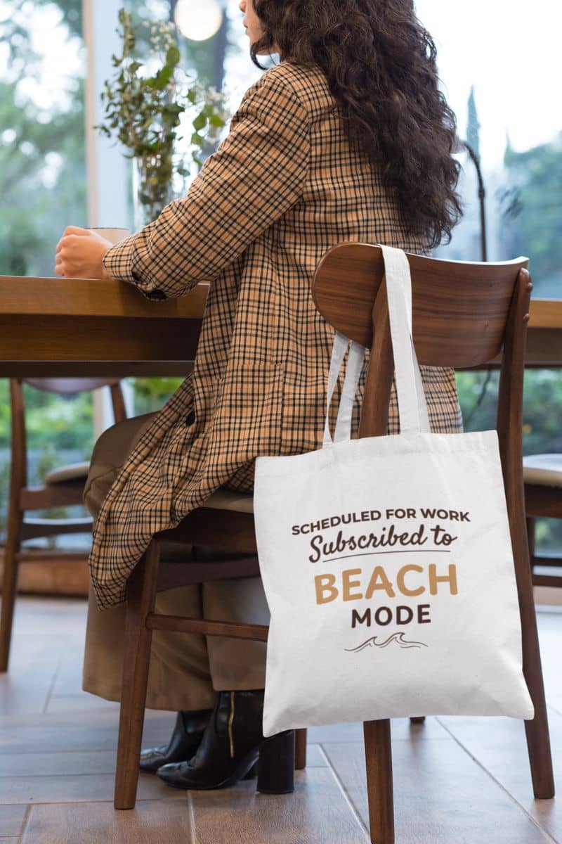 vacation tote bag with a person sitting at a table with a tote bag featuring the text 'Scheduled for Work. Subscribed to Beach' about work and beach mode.