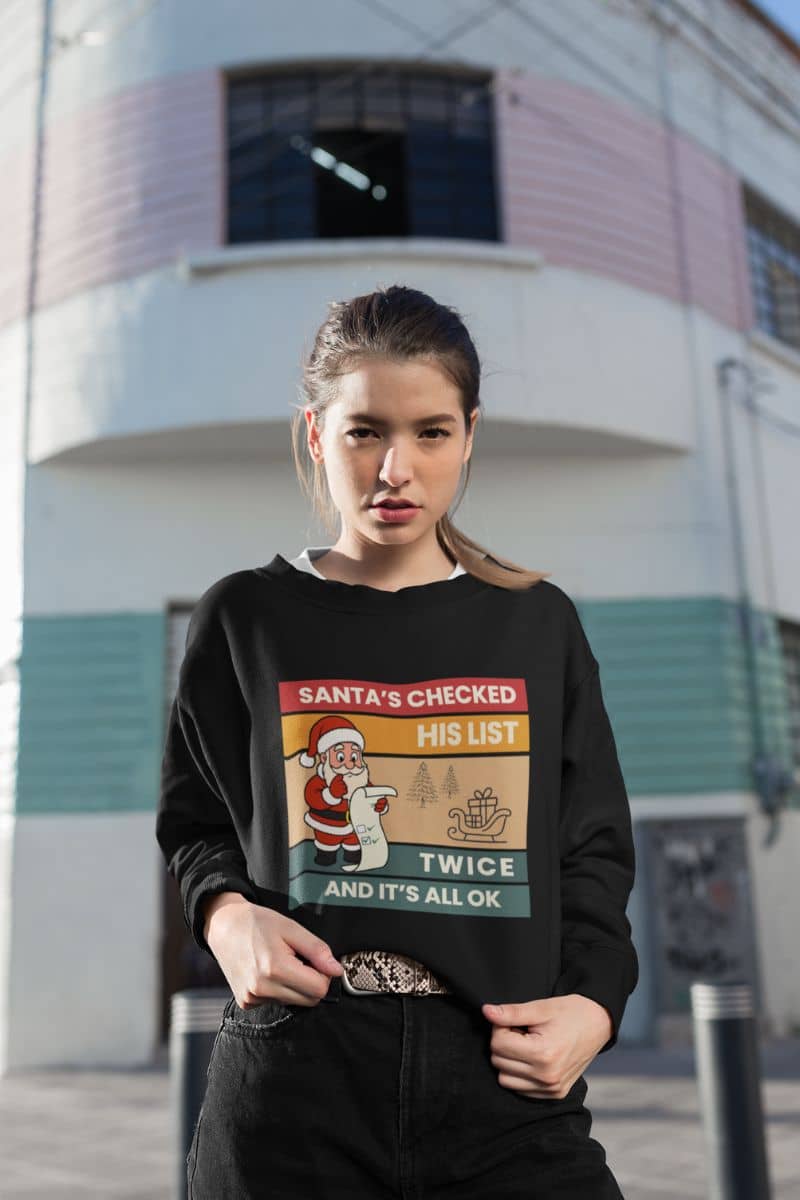 vintage christmas sweater with a person wearing a black sweatshirt with text 'Santa's Checked His List Twice And It's All Ok' in front of a building.