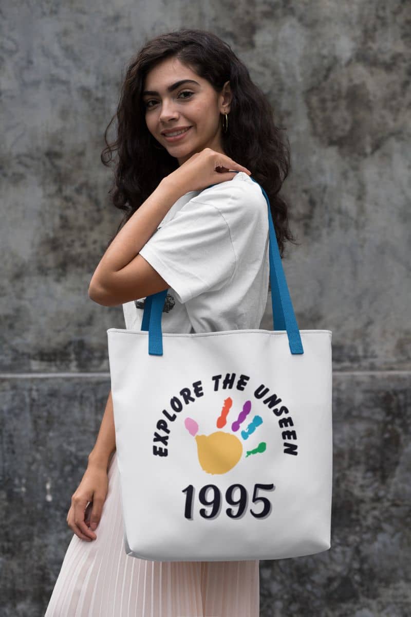 wanderlust tote bag with a woman holding a tote bag with 'Explore the Unseen 1995' design against a gray wall.