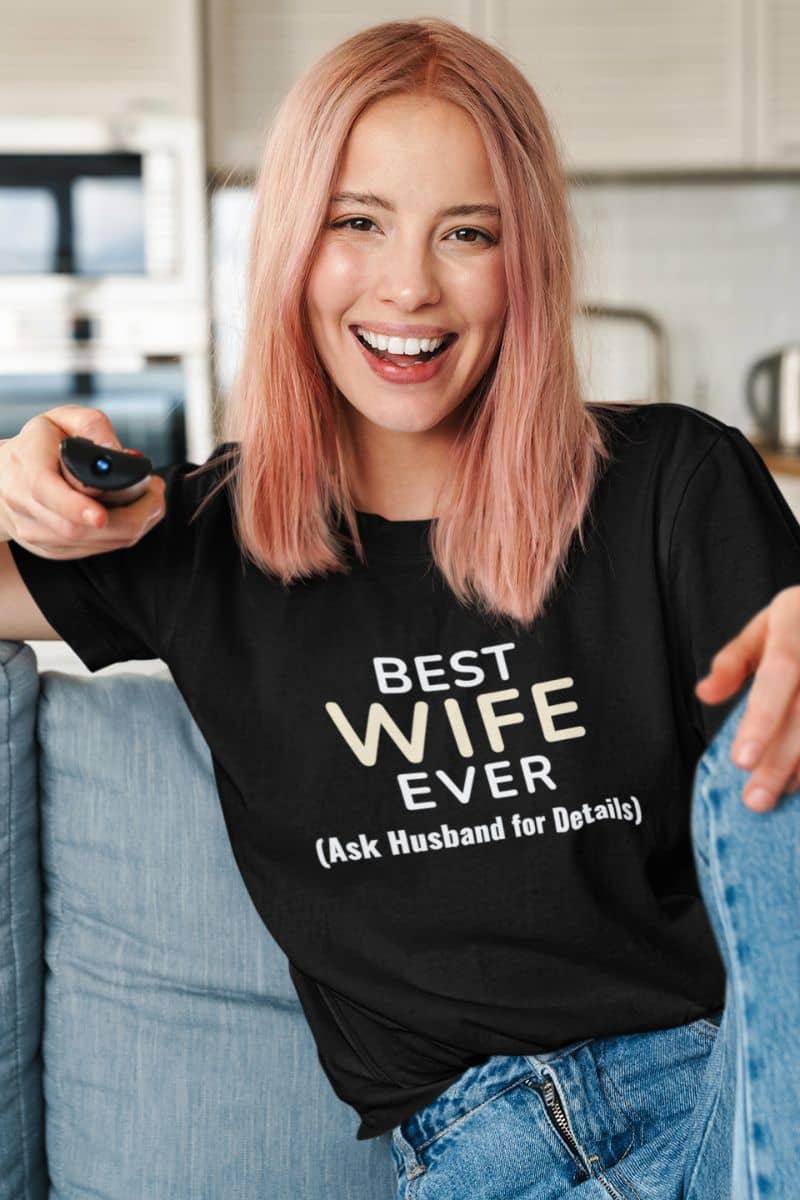 wife humor shirt with a woman wearing a black t-shirt with 'Best Wife Ever (Ask Husband for Details)' text, sitting on a couch.