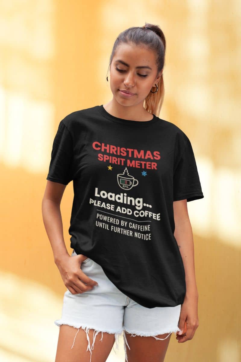 winter coffee shirt with a woman wearing a black t-shirt with 'Christmas Spirit Meter, Loading…37%. Please add coffee. Powered by caffeine until further notice' design on a blurred yellow background