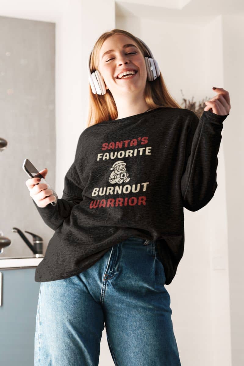 winter comfort sweatshirt with a person wearing a black sweatshirt with text 'Santa’s Favorite Burnt Out Worrior', listening to music in a kitchen.