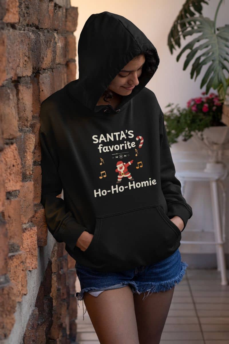 winter holiday pullover with a person wearing a black hoodie with text 'Santa's Favorite Ho-Ho-Homie' and graphics, leaning against a brick wall.