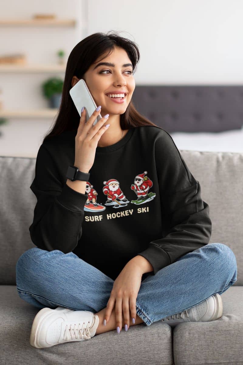 winter sports sweatshirt with a woman sitting on a couch wearing a black sweatshirt with Santa Claus designs and text, talking on a phone.