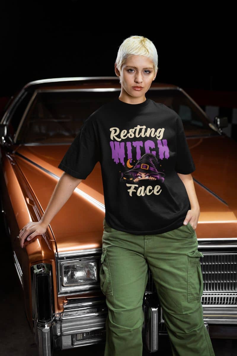 witch Halloween t shirt with a model with short blonde hair stands in front of a classic orange car, wearing a black graphic t-shirt that reads 'Resting WITCH Face' in purple and cream, paired with olive green cargo pants.