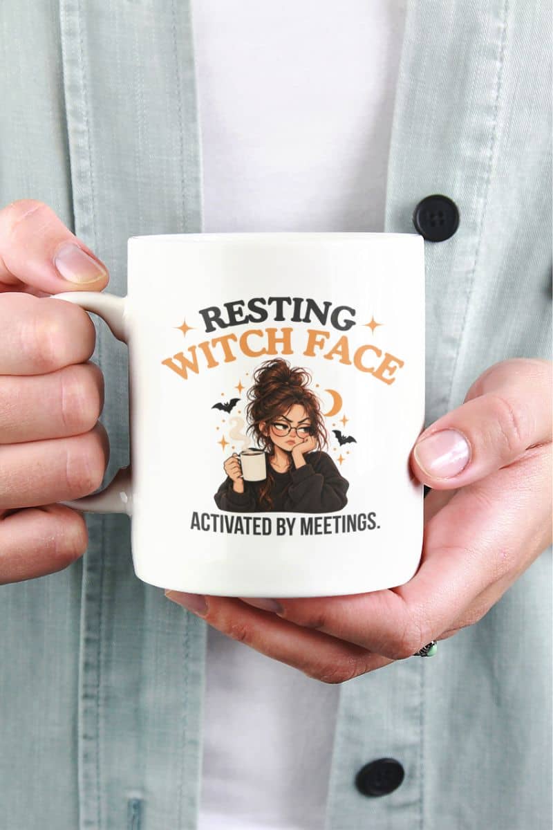 A person holding a witch mug with a Halloween-themed graphic that reads 'Resting Witch Face. Activated by Meetings.' The person is wearing a light blue button-up shirt over a white top.