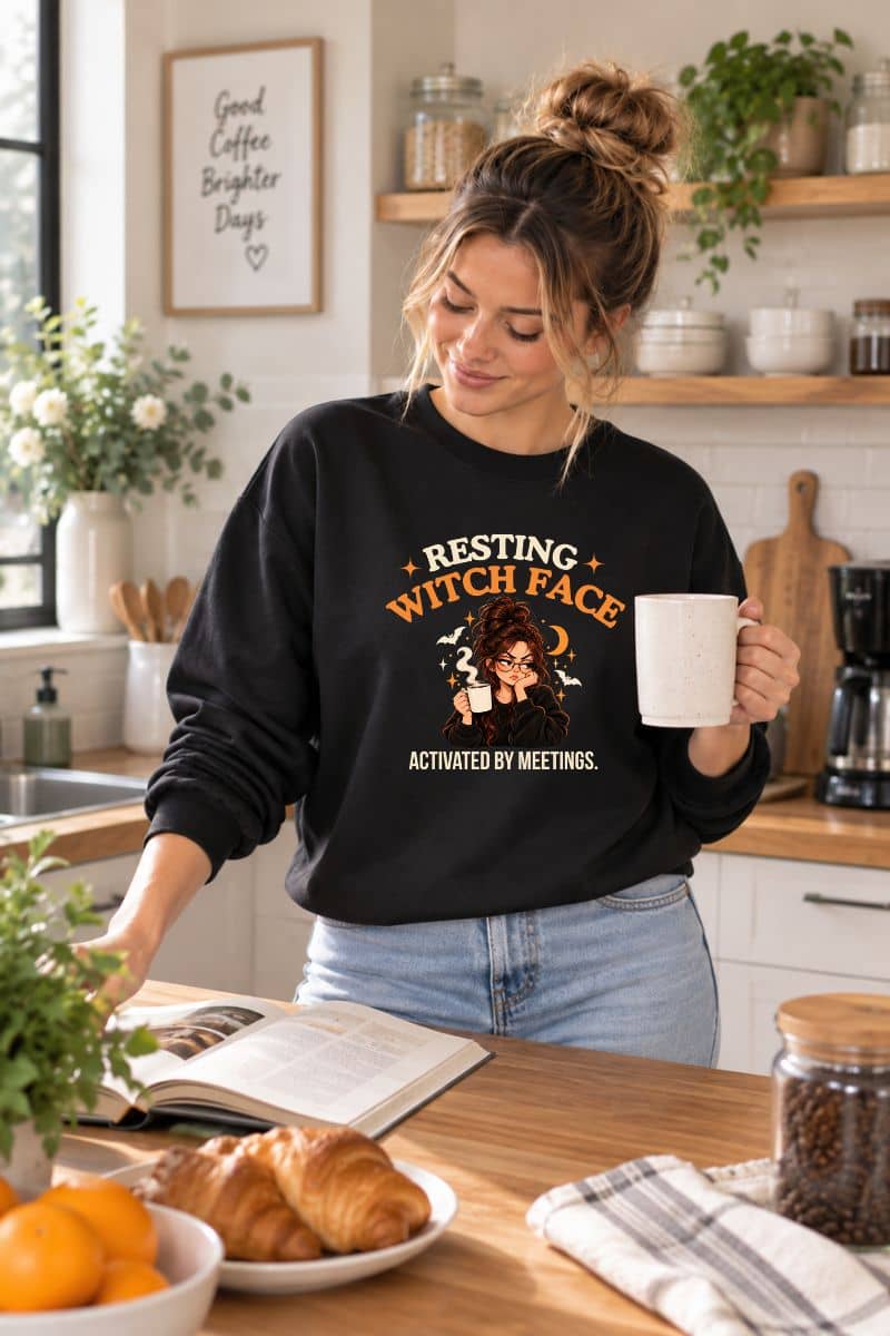 A woman standing in the kitchen, wearing a sweatshirt that reads 'Resting Witch Face. Activated by Meetings.' with a woman's face