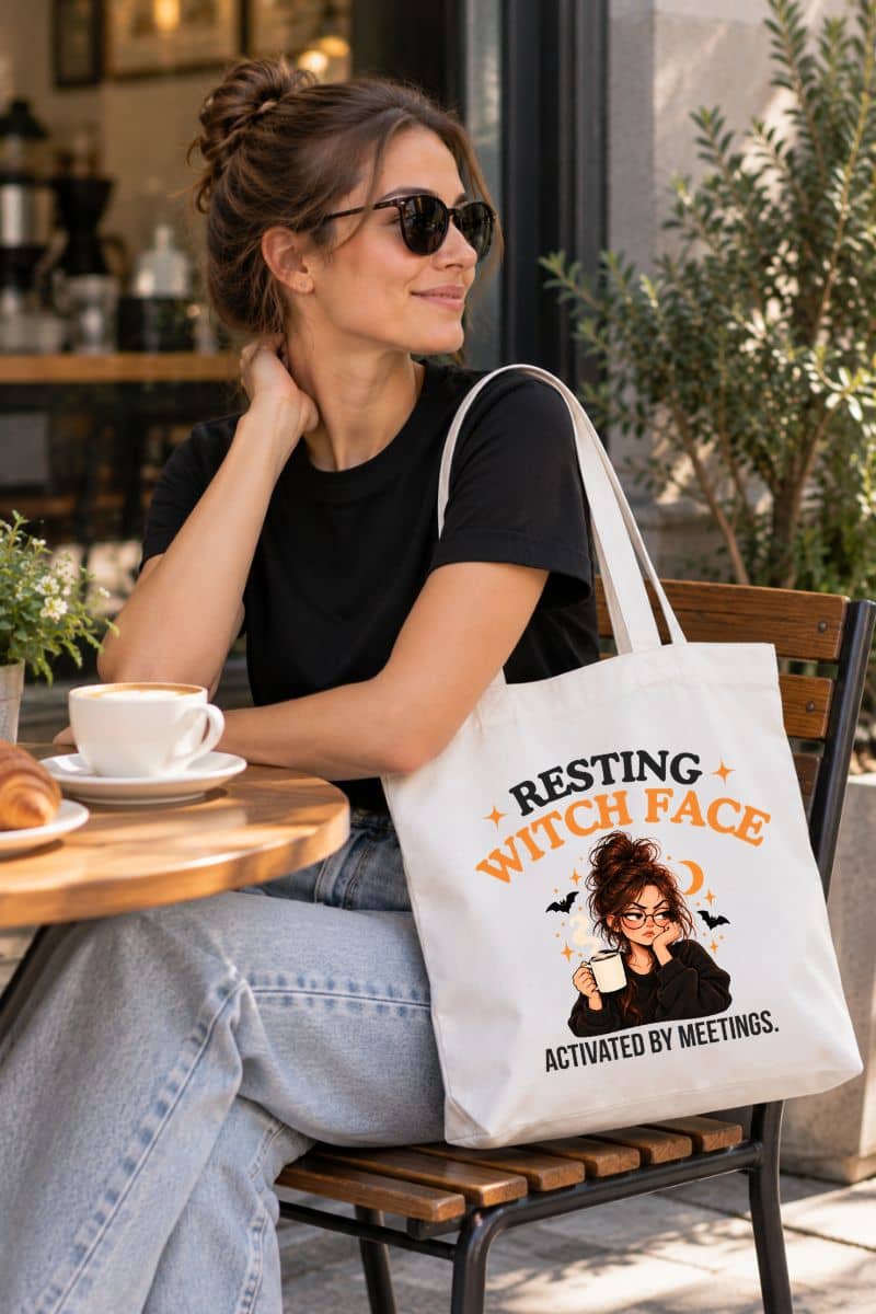 A woman sitting at a cafe table wearing a black t-shirt and jeans, with a white witch tote bag featuring a Halloween graphic and the text 'Resting Witch Face. Activated by Meetings.' resting on the bench beside her.