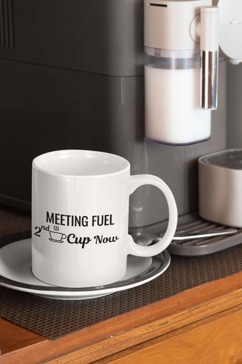 work coffee mug with a white mug with text 'Meeting Fuel: 2nd Cup Now' in front of a coffee machine.