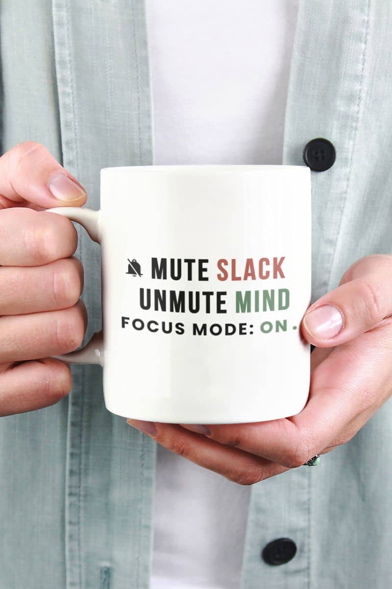 work from home mug with a person holding a mug with text 'Mute Slack, Unmute Mind, Focus Mode: On' against a light gray shirt background.