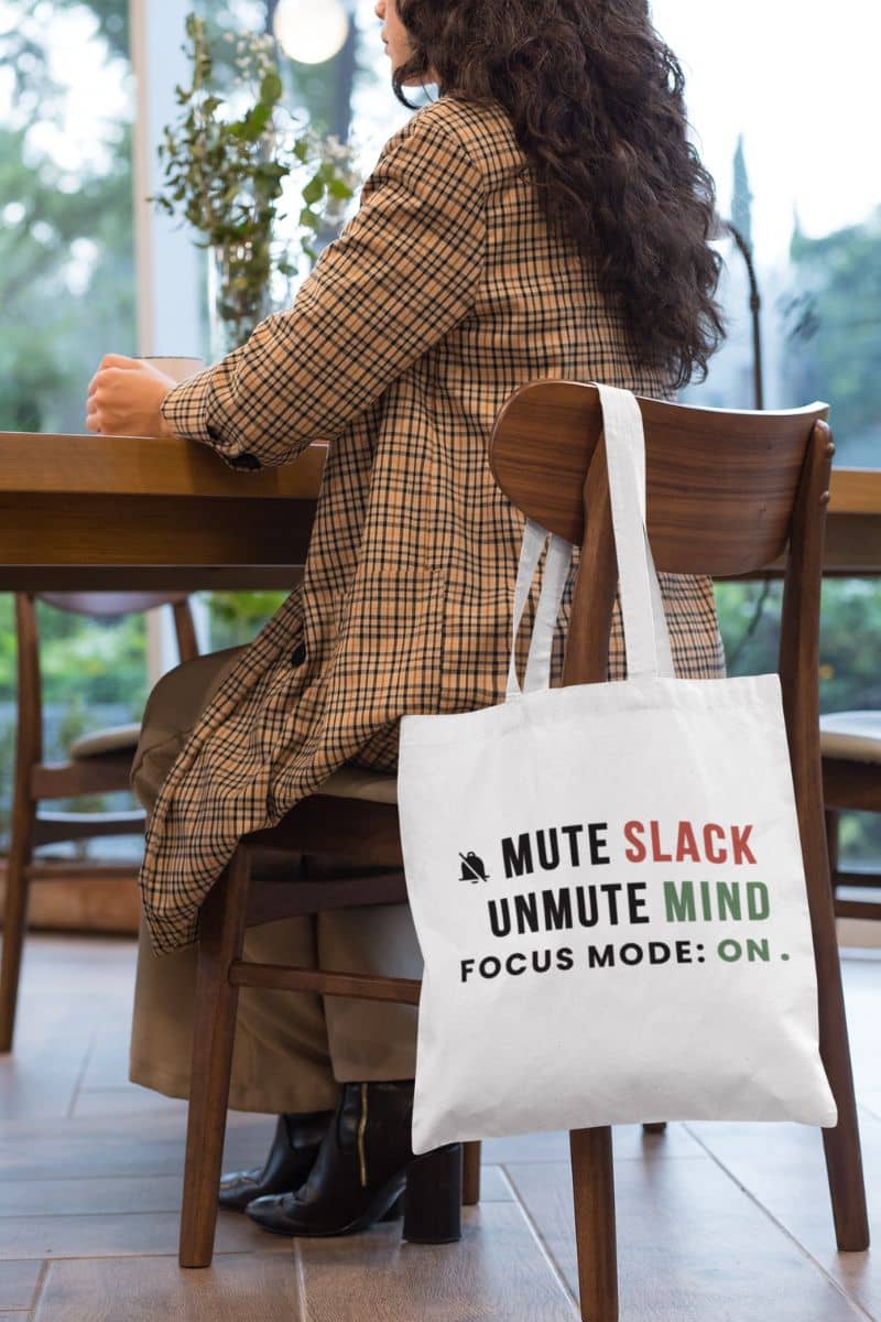 work from home tote with a person sitting at a table with a tote bag featuring text 'Mute Slack, Unmute Mind. Focus Mode: On' in a cafe.