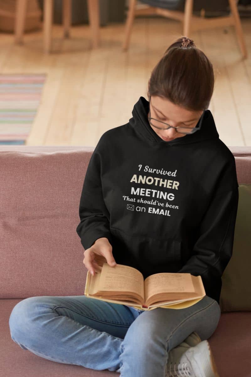 work humor hoodie with a person wearing a black hoodie with text 'I Survived Another Meeting That Should’ve Been an Email', sitting on a couch reading a book.