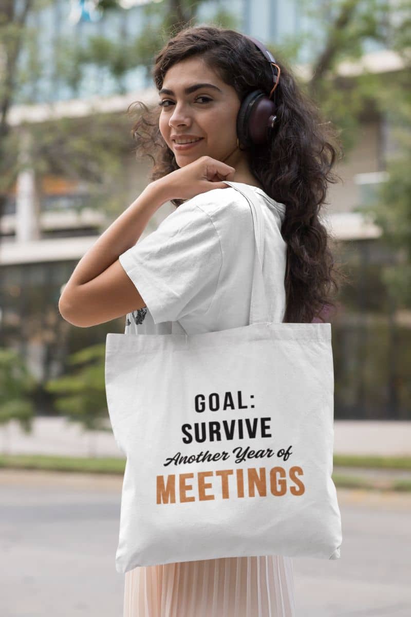 work humor tote with a woman wearing headphones and holding a tote bag with text 'Goal: Survive Another Year of Meetings', outdoors