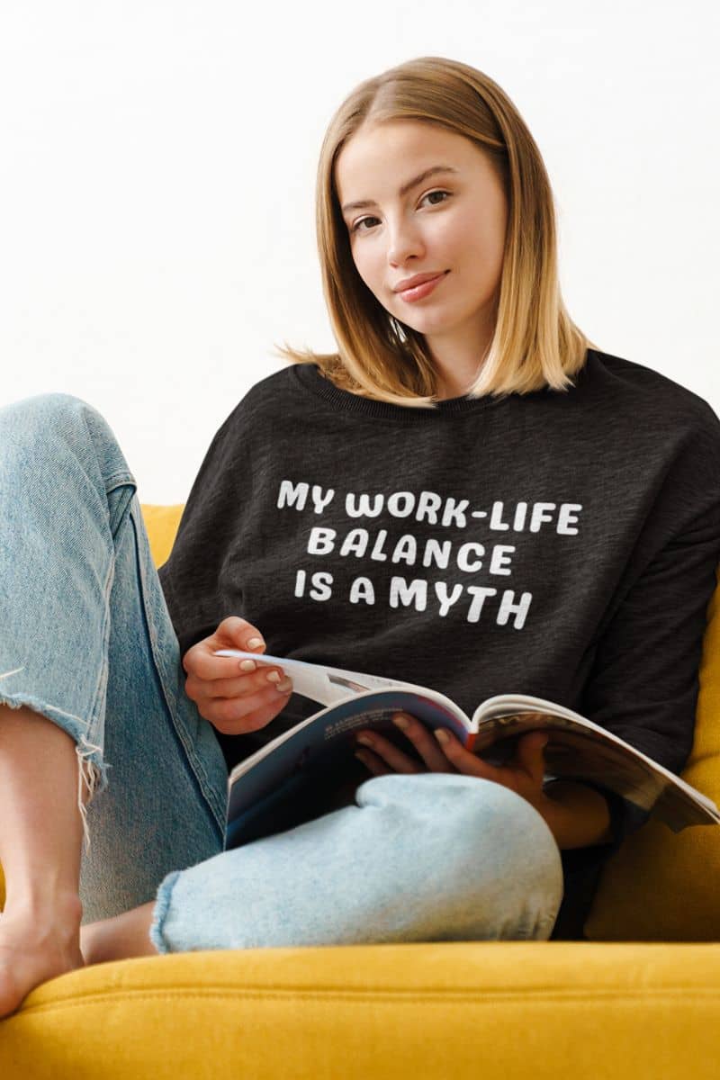 work sarcasm sweater with a woman wearing a black sweatshirt with 'My work-life balance is a myth' text, sitting on a yellow couch reading a book.