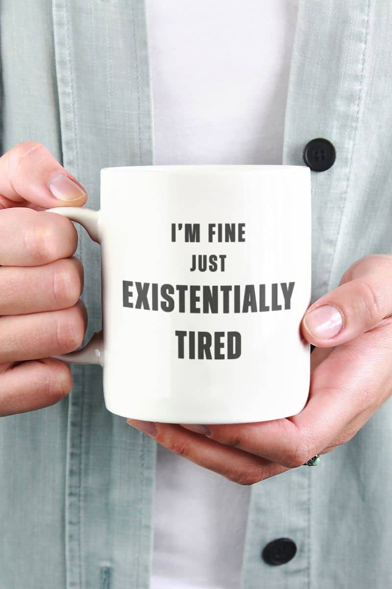 work stress coffee mug with a person holding a mug with text 'I'm fine just existentially tired' against a light gray background