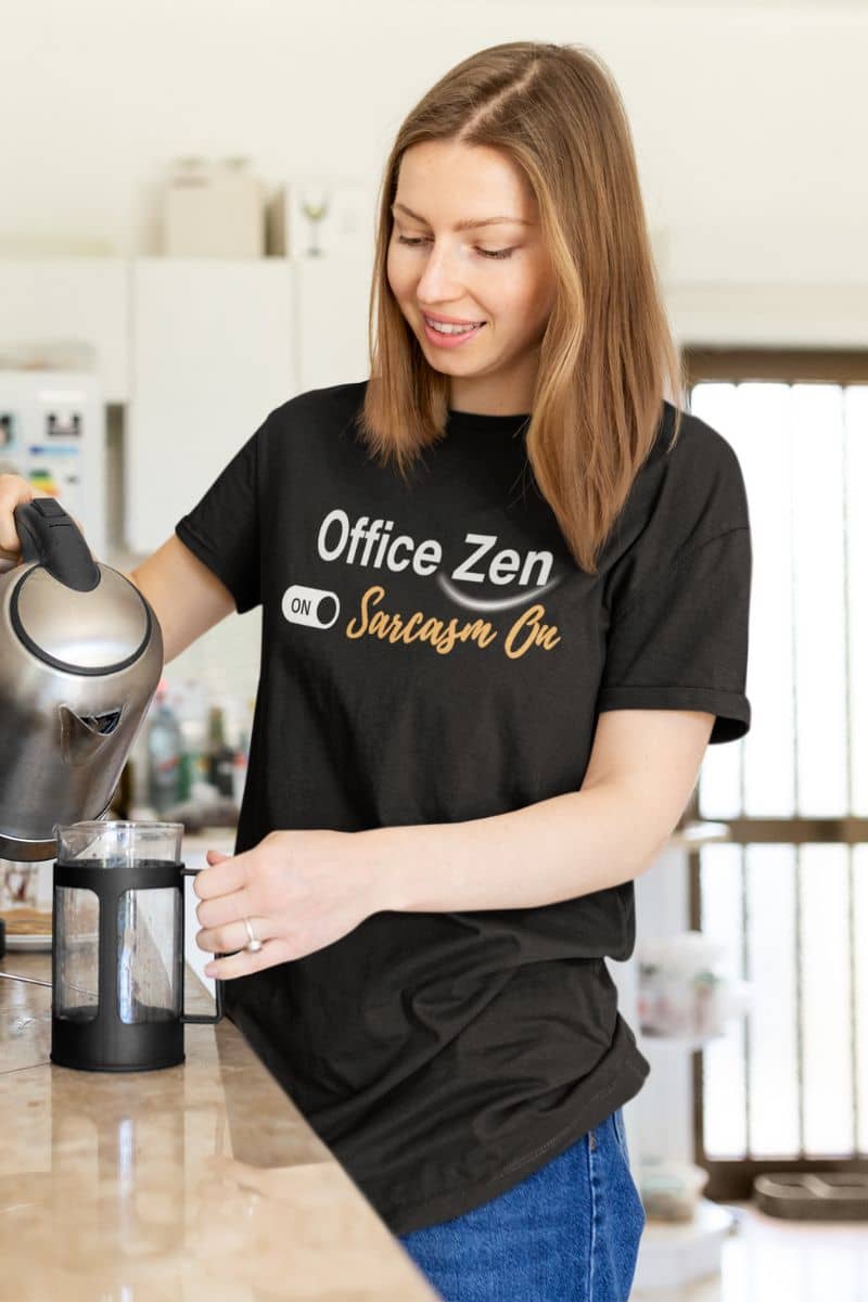 work stress humor tee with a woman in a kitchen wearing a black t-shirt with humorous text 'Office Zen, Sarcasm On', holding a kettle.
