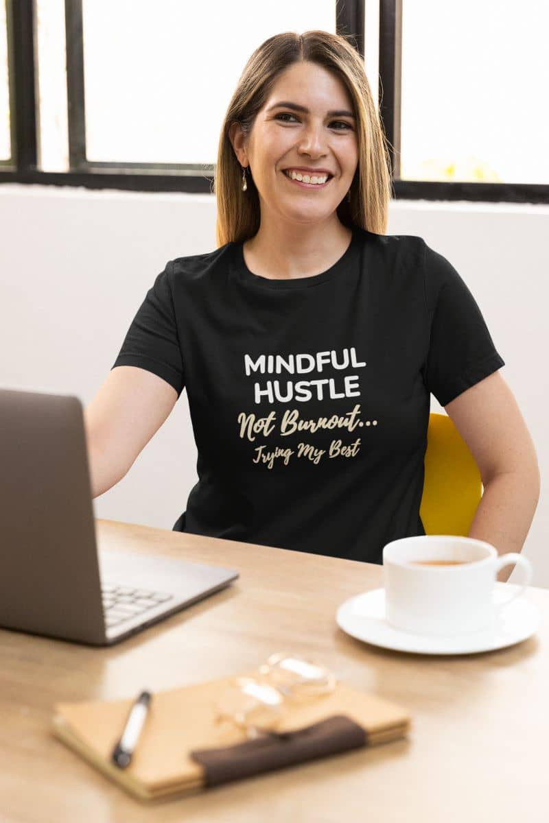work stress shirt with a woman wearing a black t-shirt with text 'Mindful Hustle – Not Burnout… Trying My Best', sitting at a desk with a laptop and coffee.