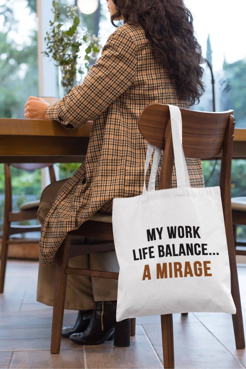 work life balance tote with a person sitting at a table with a tote bag that says 'My work life balance... a mirage' in a casual setting.