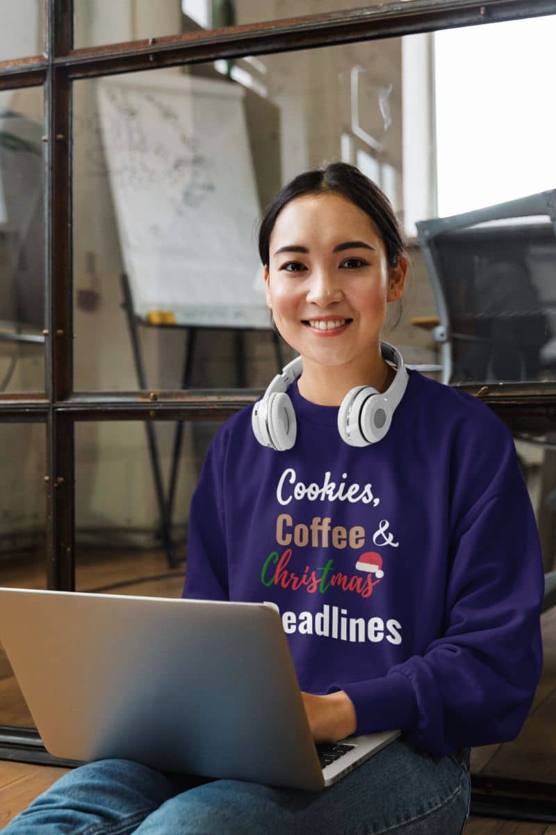 workaholic sweatshirt with a person wearing a purple sweatshirt with text 'Cookies, Coffee & Christmas Deadlines', sitting with a laptop in an office setting.