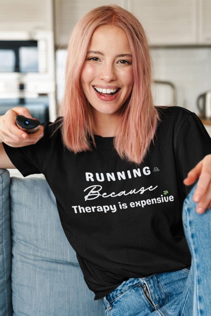 Workout humor tee worn by a young woman watching a TV show, featuring the text “Running because therapy is expensive.